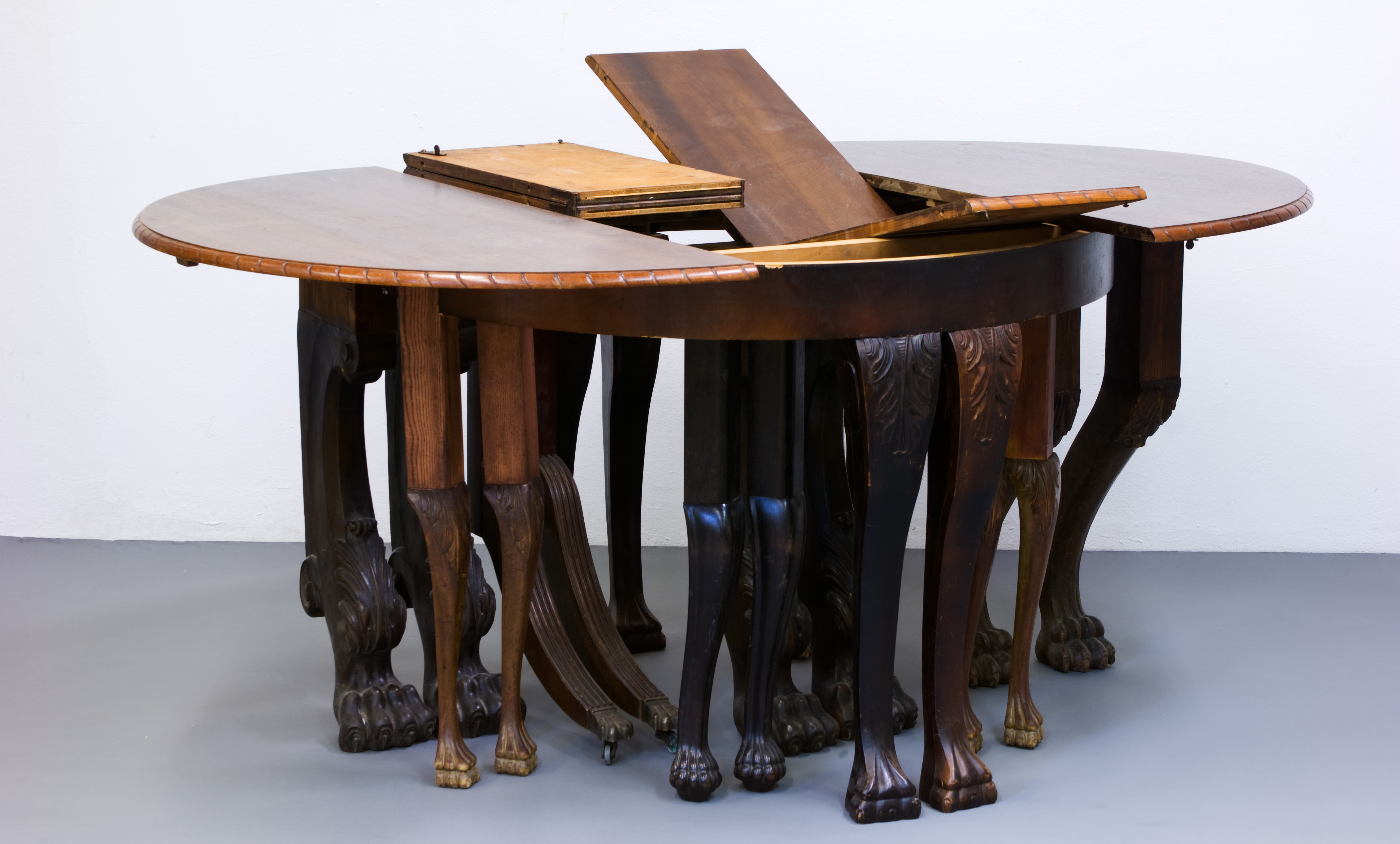 Sculpture. The two halves of a round dining room table have been pulled apart. The leaves that would expand its length sit, partially unfolded or totally folded up, in the gap. Ten pairs of dining room legs, all with clawed feet, hold the table up.