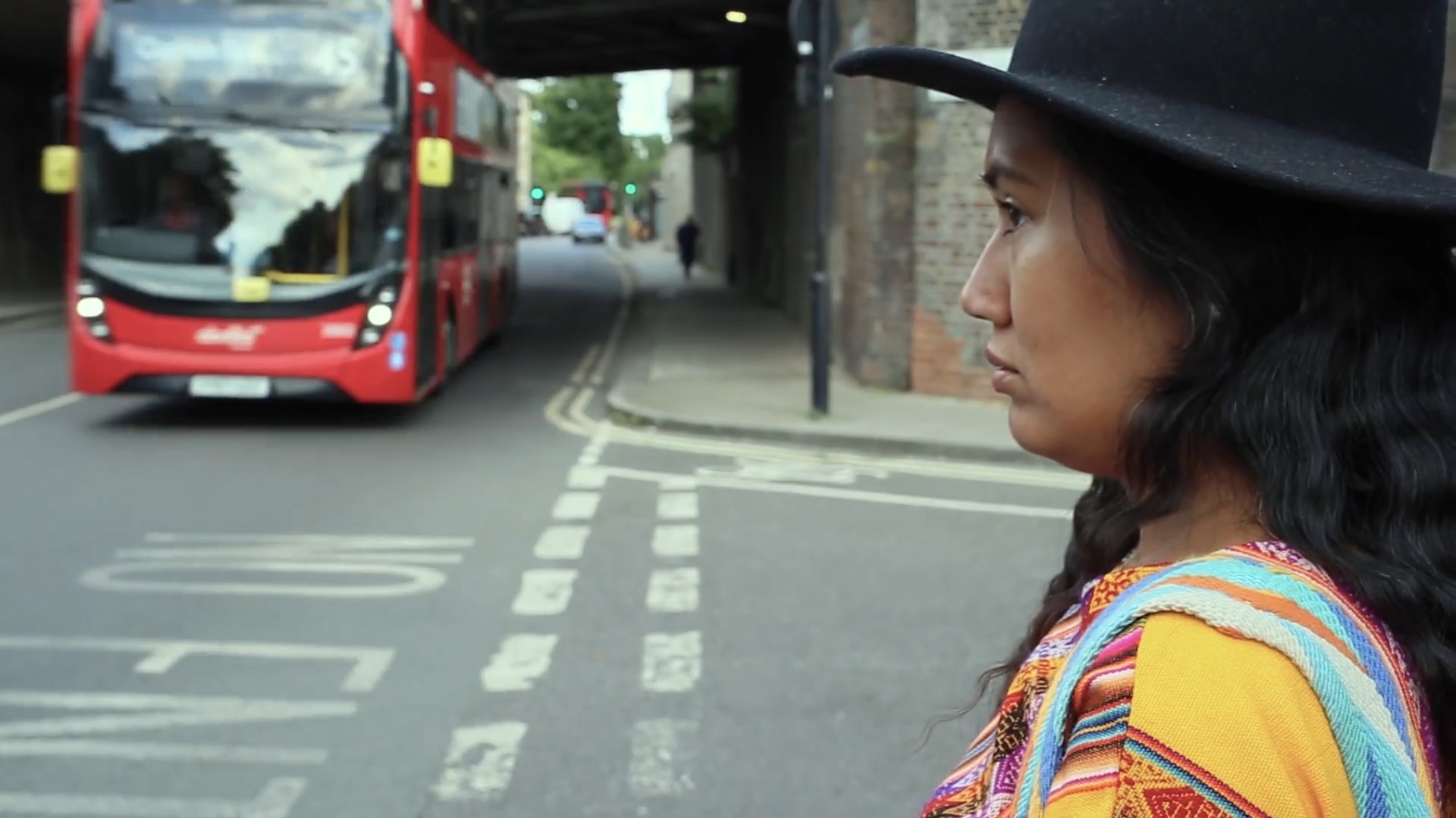 Video still. A dark-skinned person stands to our right, close to the camera, on a city street. Looking off to our left in profile, they wear a brimmed black hat over long black, wavy hair. The garment is striped with vividly coloured patterns. A red double-decker bus comes towards us down the street, off to our left.