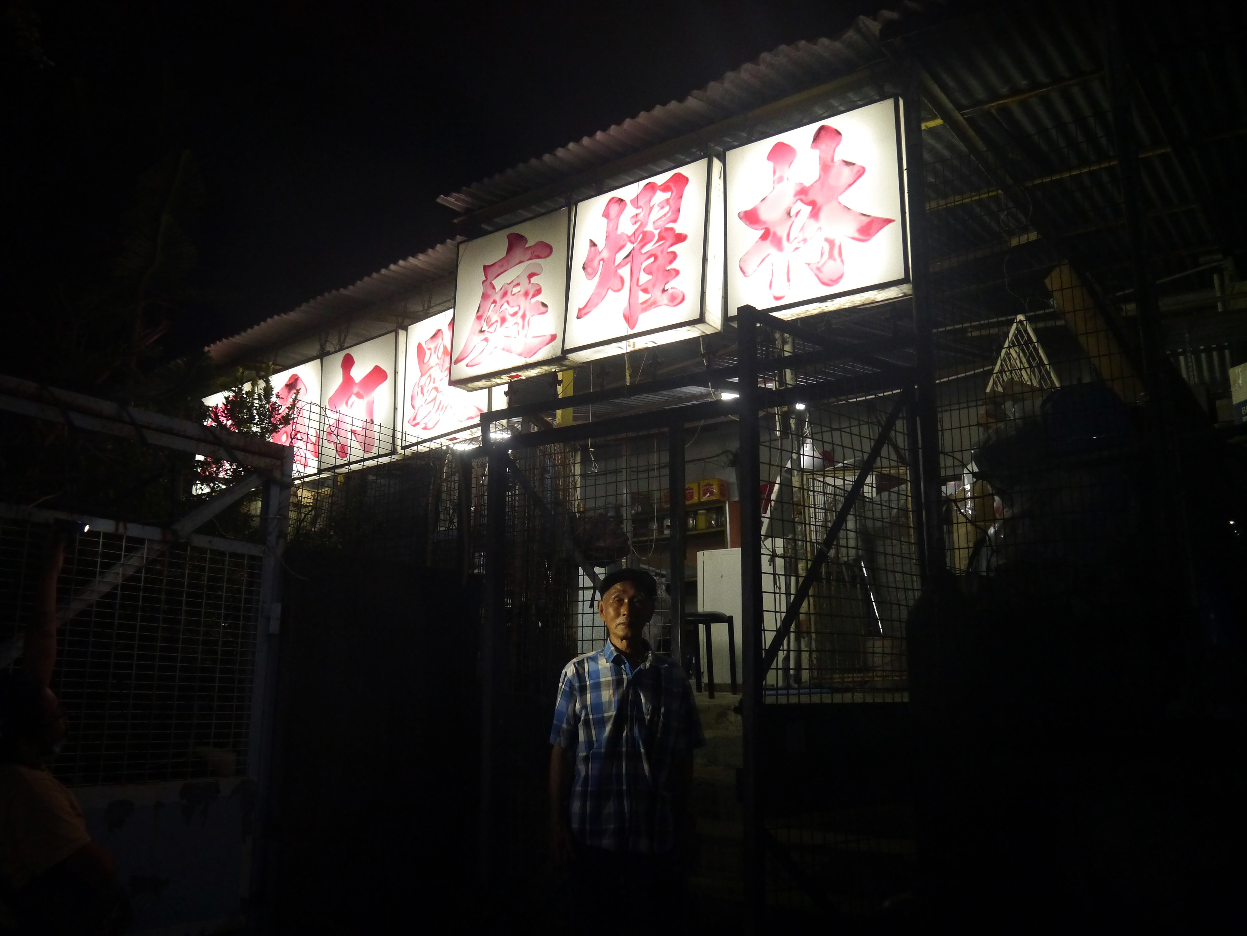 A medium-skinned man wearing a cap and a blue plaid shirt stands under an illuminated sign with red, Chinese characters under a night sky. A workshop seems to be enclosed with wire fencing under the sign.