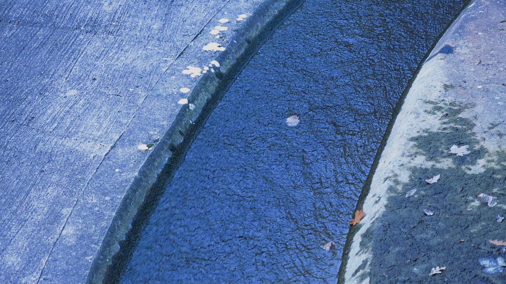 Video still. We look straight down onto what appears to be a slightly curving canal or aqueduct filled with lapis-blue water. Cement embankments line each side in paler shades of blue.