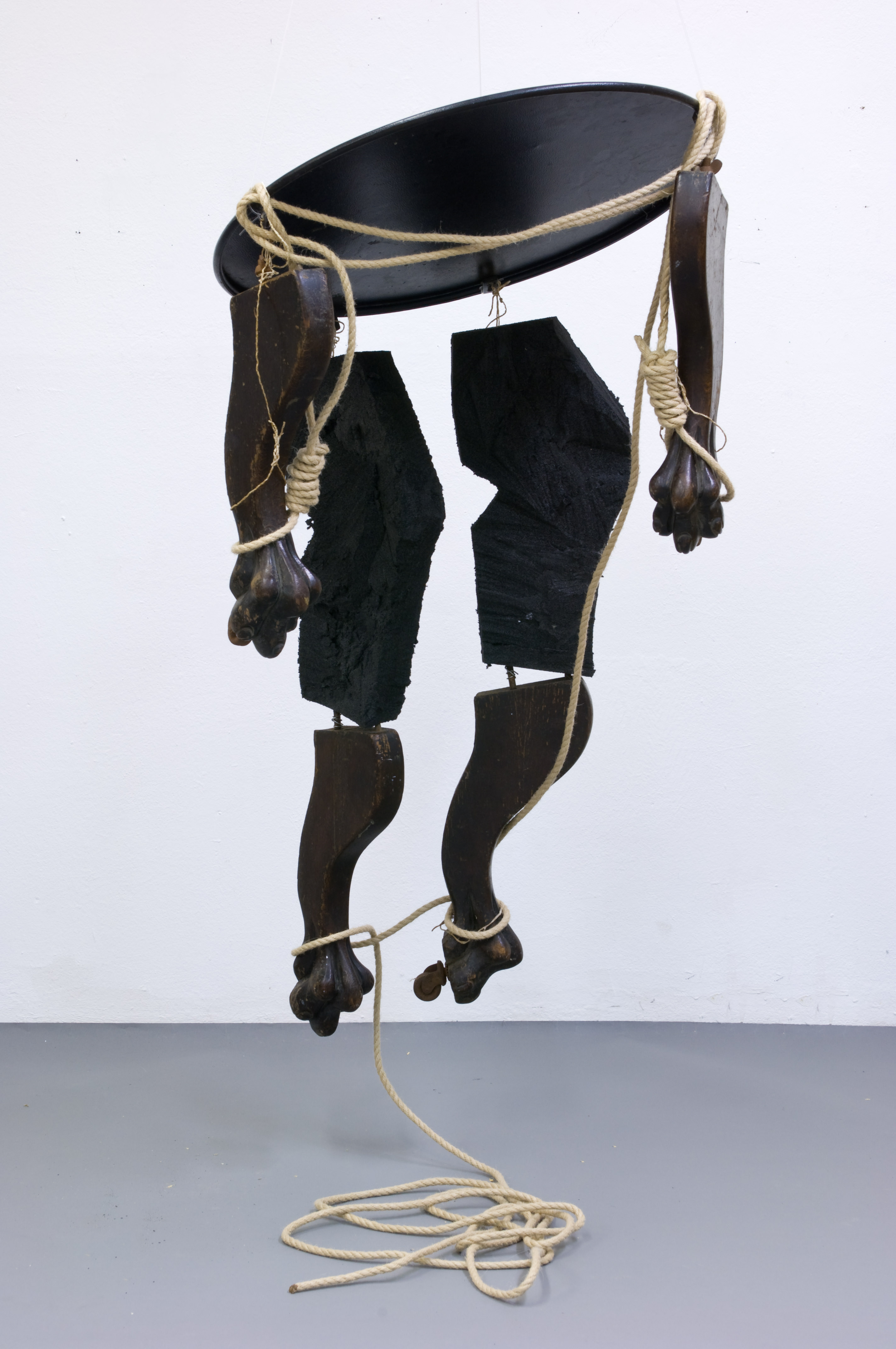 Sculpture. The round tabletop and four legs, each ending with a clawed foot, are cut apart and reassembled with wire and hung like a puppet or mobile to resemble a human body. Nooses encircle what would be the wrists, and other parts are wrapped with rope.