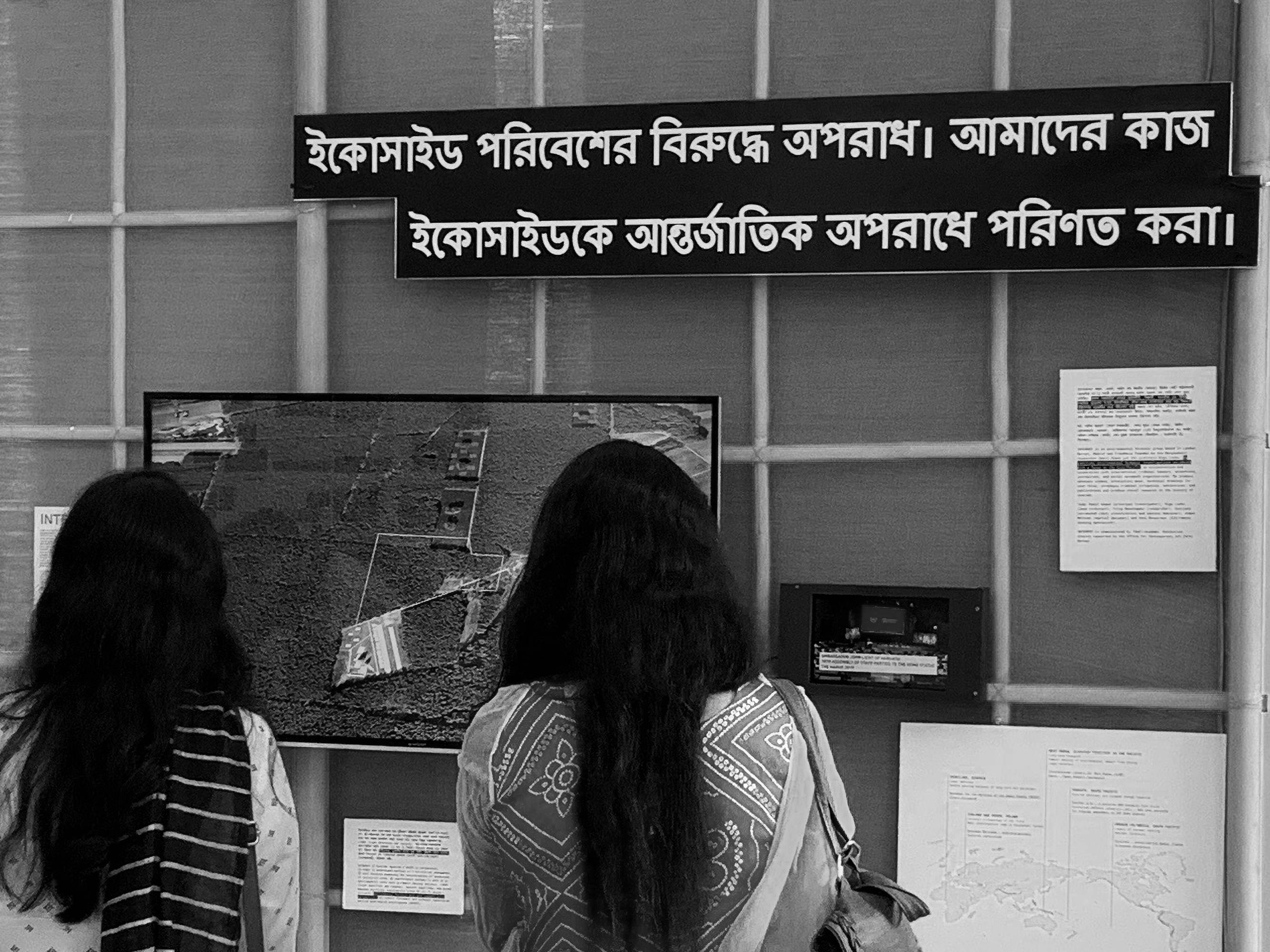 Installation. Black and white photograph shows two people with long black hair looking away from us, at a map and texts arranged on a wall.