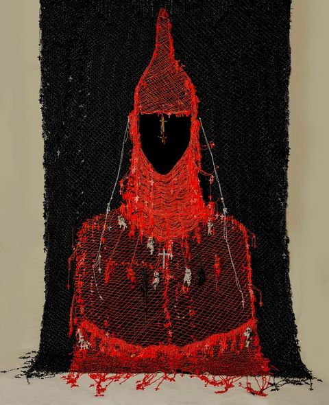 Mixed media. The red-hooded, red-caped form of a person seems drawn in thread against a black background. Small crosses and statuettes hang from the cowl and robe. The area where the face would be is flat black.