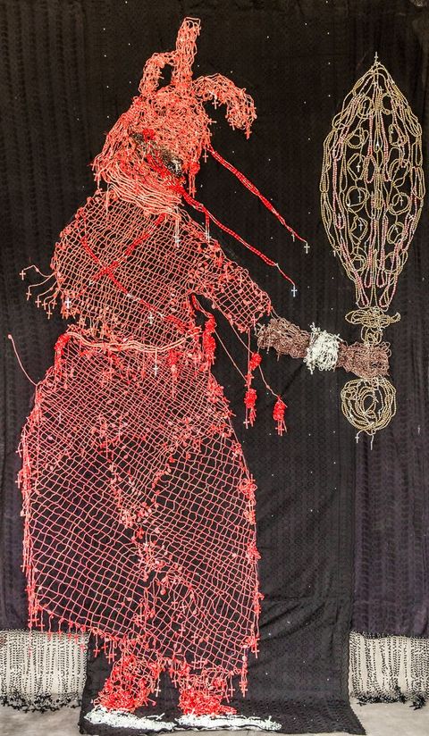 Mixed media. A red silhouette of a person wearing a hood and robe and holding out a sword or other object seems drawn in red and gold against a black background. The person’s arm is brown, and the wrist has a white or silver cuff bracelet.