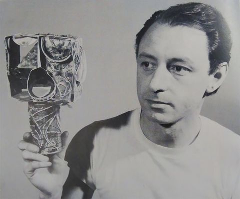 Black and white photograph. Shown from the chest up, aa pale-skinned man with his dark hair brushed back from his forehead and wearing a white t-shirt holds a sculpture in one hand. He looks to our left with dark eyes past the sculpture, which is a cube on top of a cylinder. The abstracted head, the cube, has exaggerated eyes and a wide-open mouth. The entire work is collaged.
