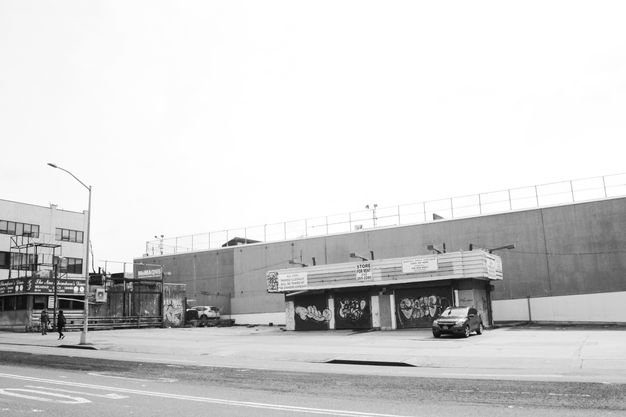 Black and white photograph. One car is parked in front of a long, warehouse-like building.