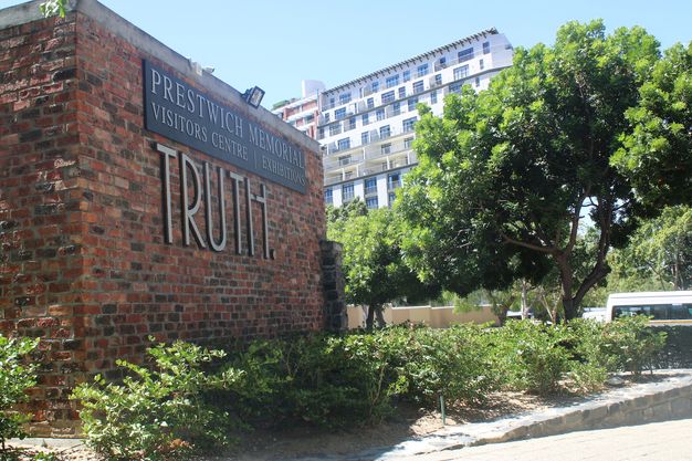 A brick wall with tall capital letters reading “Truth” is close to us, in front of a high-rise building.