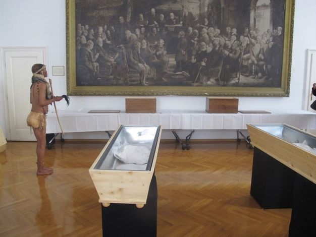 Two coffins lie in a room hung with a black and white work of art showing a gathering of men. Two wooden boxes sit on a table in under the artwork. To our left, a medium-skinned person wearing a collar, headband, and cloth around his hips addresses the boxes.