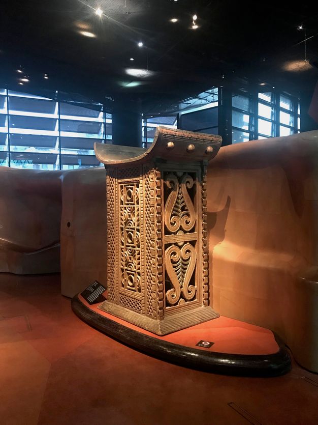 A rounded seat is atop an elaborately pierced and carved rectangular base. The set curves like a shallow letter C. The object is spotlit in a museum with windows lining the far wall.