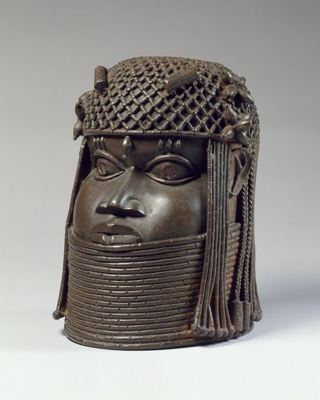 Sculpture. Freestanding brass sculpture of a head with stylized features. The crown of the head is covered with a net. Ornaments hang down the sides of the face and along the forehead. The face has round cheeks and wide-open eyes. A tall choker comes up to just below the lower lip.