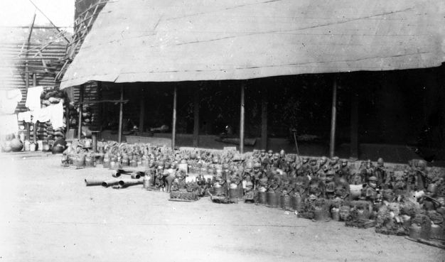 Black and white photograph. Dozens, maybe hundreds of statuettes and objects are lined up in rows in front of a building with a long, sloping roof.