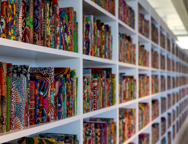 Installation. White shelves are filled with colourfully patterned books. We see the installation from close to the shelves, so they extend into the distance out of focus as far as we can see.