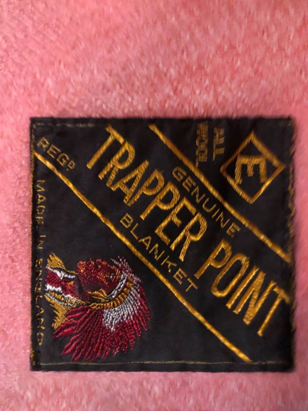 Label. A fabric label stitched to a pink blanket is a black square with the brand stitched in gold. A dark-skinned head wearing a feathered headdress is embroidered over text reading, “Made in England”.