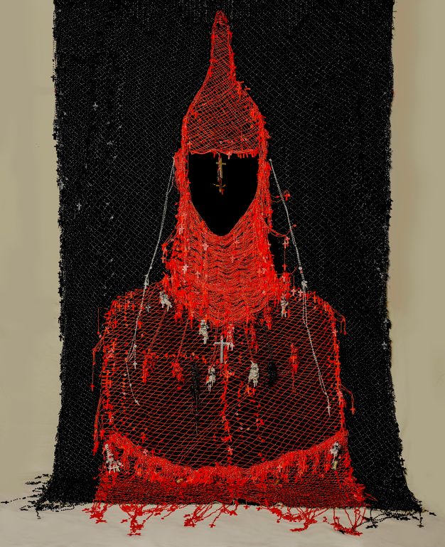 Mixed media. The red-hooded, red-caped form of a person seems drawn in thread against a black background. Small crosses and statuettes hang from the cowl and robe. The area where the face would be is flat black.