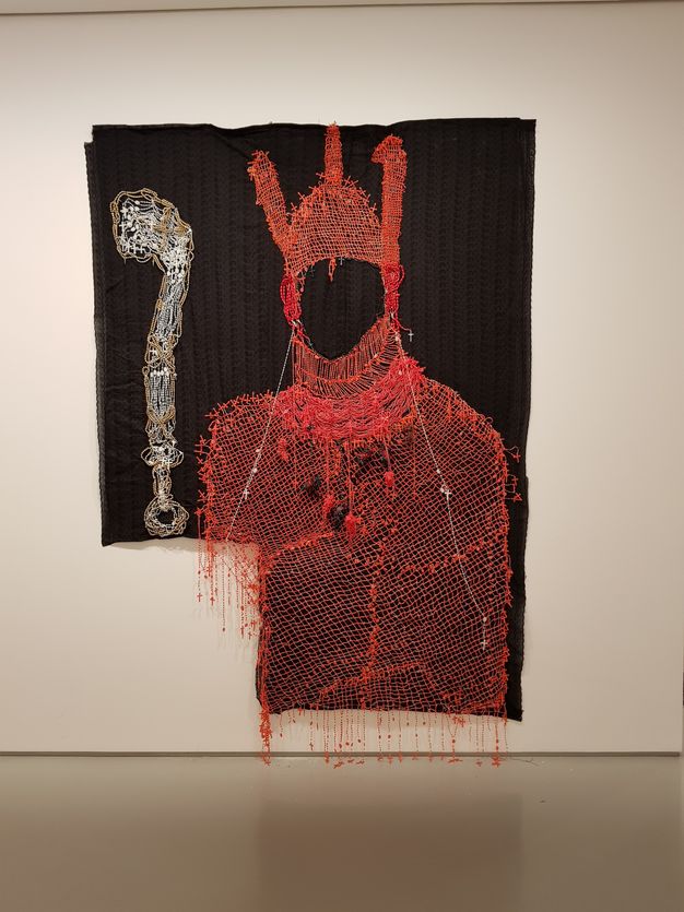 Mixed media. A red silhouette of a costume seems drawn in thread against a black background. The hood has three vertical projections. Tiny red crosses are hung all over the costume, especially around the edges. A silvery staff is outlined in gold to our left.