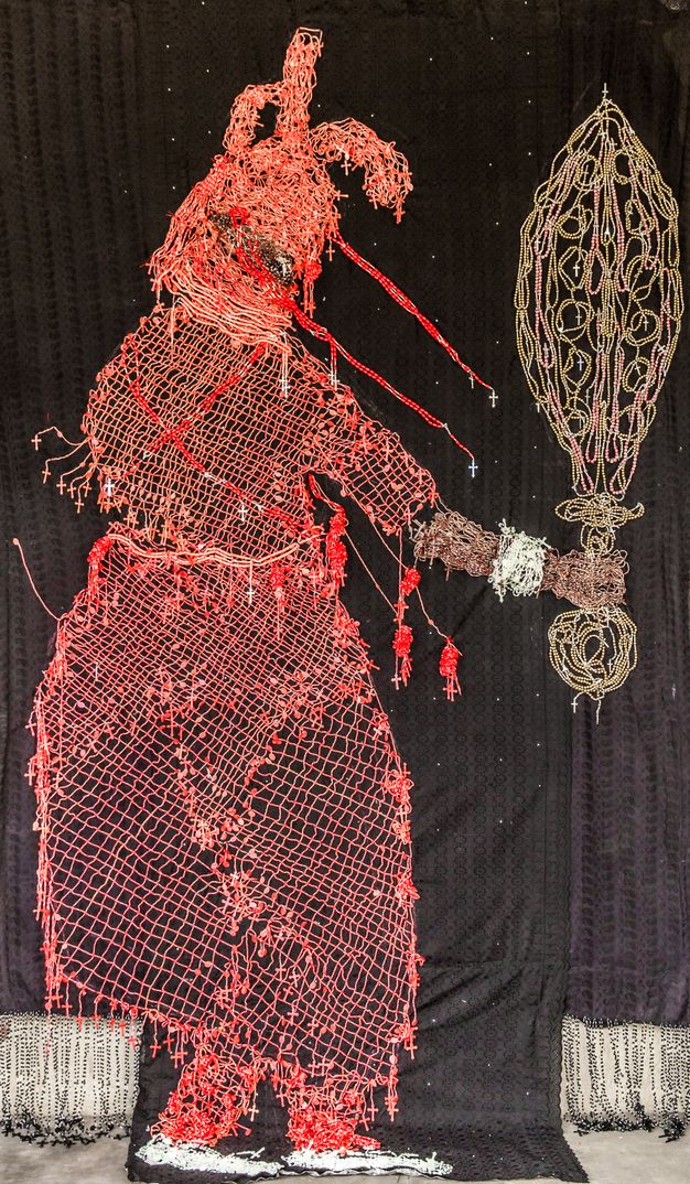 Mixed media. A red silhouette of a person wearing a hood and robe and holding out a sword or other object seems drawn in red and gold against a black background. The person’s arm is brown, and the wrist has a white or silver cuff bracelet.