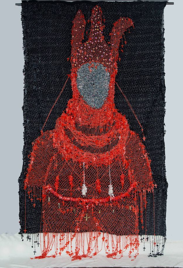 Mixed media. A red silhouette of a costume seems drawn in thread against a black background. The hood has three vertical projections. Tiny red crosses are hung all over the costume, especially around the edges. The space where the face would be is silver.