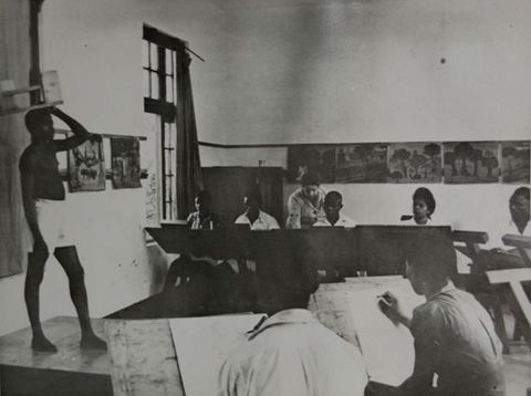Black and white photograph. A model with dark skin, wearing white shorts, stands with one hand on his head in front of a class. Seven students, all with dark skin, are visible in this image. One is a woman and six appear to be men. A lighter-skinned woman leans over the shoulder of a student across the room from us.