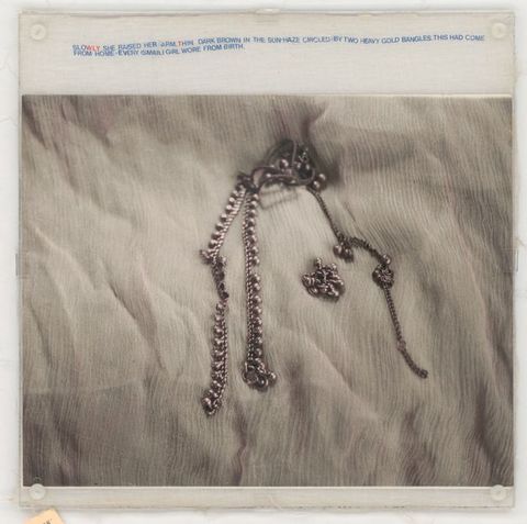 Mixed media. A black and white photograph shows a piece of silver jewellery lying on a white, wrinkled cloth. Mostly blue text in all caps printed on muslin above the image reads, “Slowly she raised her arm, thin dark brown in the sun-haze circled by two heavy gold bangles. This had come from home – every Ismaili girl wore from birth”. The WLY in “slowly” and T in “thin” are red.