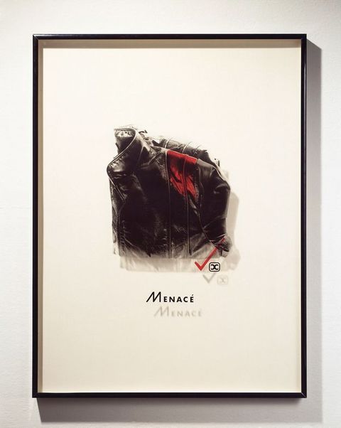 Mixed media. A photograph of a high-collared, folded, black leather jacket has been printed onto a piece of Plexiglass and mounted in a frame so it casts a shadow on the mat behind it. A swipe of red has been added to the front shoulder of the jacket. The word “Menacé” is below.
