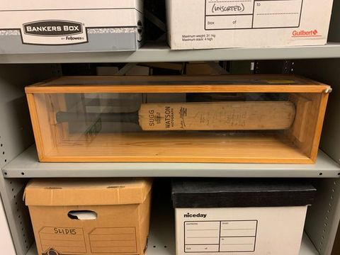 Cricket bat. On a metal storage shelf, a cricket bat is enclosed in a wood and glass case. The manufacturer’s information is near the black handle, reading “Sugg and “Watson Autograph”. A column of about a dozen handwritten signatures along the length of the bat can barely be discerned.
