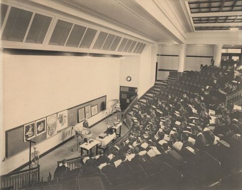 Black and white photograph. From a back row off to one side, we look down at about twelve rows of seats curving around a long blackboard hung with illustrations and diagrams in this lecture hall. A man stands between the blackboard and tables lined with specimens and materials. Students fill the seats down the centre of the hall.