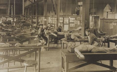 Black and white photograph. At least a dozen dead, human bodies in various stages of decay are laid out on tables. Three men cluster around one body to our right. Two more examine other bodies nearby. The room has a high ceiling and walls are hung with illustrations and diagrams.
