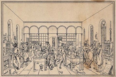 Print. Shown in black lines on light brown paper, men stand or sit at two long tables piled with equipment. Shelves also line the walls, and a few bits of paper and other detritus are scattered on the floor.