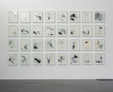 Installation photograph. Grey and white drawings in white frames are hung in a grid of eight rows by four columns. Landmasses silhouetted in grey or black are layered. Some compositions are nearly full and others sparse.
