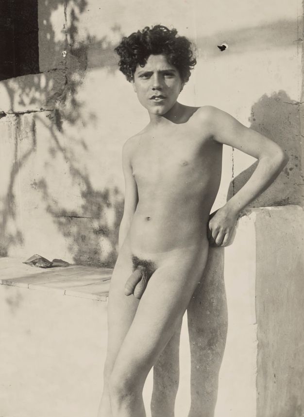 Black and white photograph. Shown from the knees up, a nude boy stands leaning against a ledge in front of a white, exterior wall. He has curly, dark hair, and his full lips are open over a cleft chin. His genitals stand out against his smooth, pale skin.