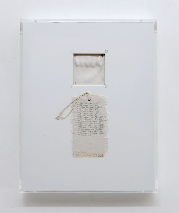 Mixed media. A square window is cut near the top centre of a rectangular white card to reveal what might be a plaster cast of a baby’s hand. Words are typewritten on a swatch of cotton fabric below. The fabric is affixed to a tag with twine strung through a hole at the top.