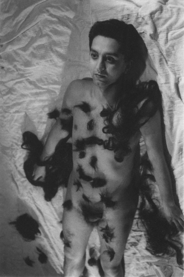 Black and white photograph. A nude man lies on a sheet, his body covered in tufts of black fur or hair. Longer tresses are around one shoulder and over or near both hands. We look down at him so he could as easily be standing, back and arms pressed to a wall.