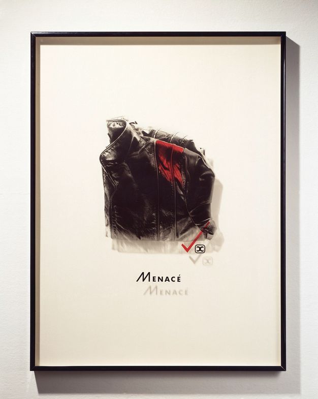 Mixed media. A photograph of a high-collared, folded, black leather jacket has been printed onto a piece of Plexiglass and mounted in a frame so it casts a shadow on the mat behind it. A swipe of red has been added to the front shoulder of the jacket. The word “Menacé” is below.