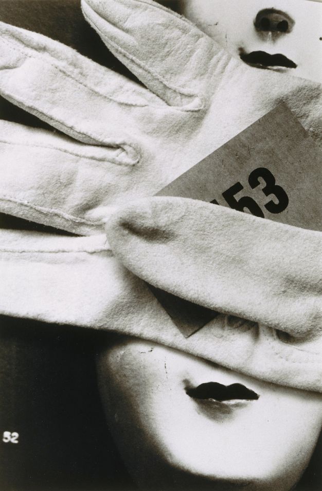 Black and white photograph. An empty white glove has a piece of paper printed with the number “53” tucked under the thumb. The glove fills the top two-thirds of the picture. Two pale doll’s faces with dark, smiling lips are seen from the nose down. There is one in the upper right corner and one in the corner below.