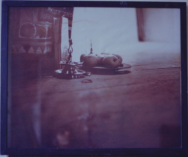 Colour photograph. In tones of violet and smoky purple, we look down onto a wooden tray holding a teapot, mug, and two more vessels, one with a spoon. The carpet beneath is woven with a stylized, floral design. The toes of one foot creep up over the bottom edge of the tray, and the other foot stands next to it. The photograph is in a purple frame.