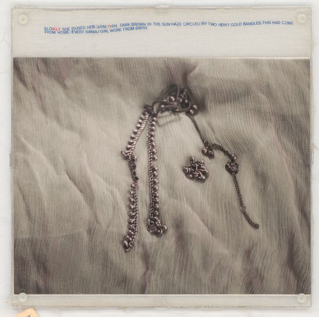 Mixed media. A black and white photograph shows a piece of silver jewellery lying on a white, wrinkled cloth. Mostly blue text in all caps printed on muslin above the image reads, “Slowly she raised her arm, thin dark brown in the sun-haze circled by two heavy gold bangles. This had come from home – every Ismaili girl wore from birth”. The WLY in “slowly” and T in “thin” are red.