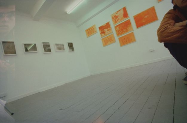 Installation photograph. From low to the grey-painted wood floor, we look into a corner of a gallery hung with black and white photographs on the left wall and orange-drenched photographs to our right. Close to us, to our right, is the elbow and knee of a person sitting sits with elbows on knees and ankles crossed.