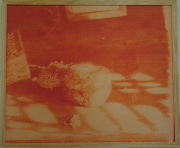 Colour photograph. In tones of orange, objects on a floor are difficult to make out but seem to include a decorative box and an embroidered object. The photograph is in a blonde wood frame.