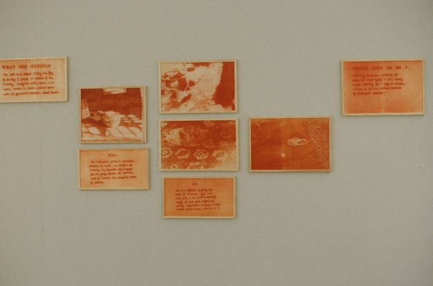 Installation photograph. Eight framed pictures or text, all seeped in tones of orange, are hung in uneven columns. Four photographs show objects on carpeted or wood floors. The other four works are handwritten words. The text is difficult to make out in this image, but the title is legible on the top left frame.