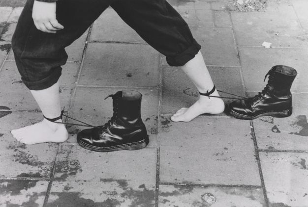 Film still. Shown from the hips down, a barefooted person walks on a sidewalk in this black and white image. One foot is planted far in front of the other, and each is tied around the ankle with the long laces of distinctive high, black Doc Martens boots.