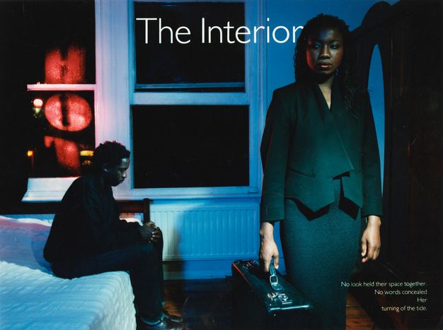 In a dimly lit room, a woman stands holding a suitcase to our right. A man sits on a bed a bit farther from us, to our left. Both have brown skin, black hair, and wear dark clothing. Red lights are reflected in the window on the far side of the bed. White text at the top centre of the composition reads, “The Interior”.