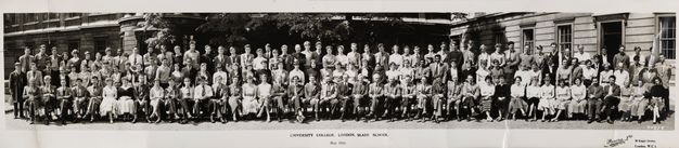Black and white photograph. Around 125 men and women stand or sit with arms crossed, by their sides, or in their laps in four long lines in this panoramic class photograph. Most have pale skin, but there are a few men with darker skin. The men mostly wear suits and ties, the women dresses.