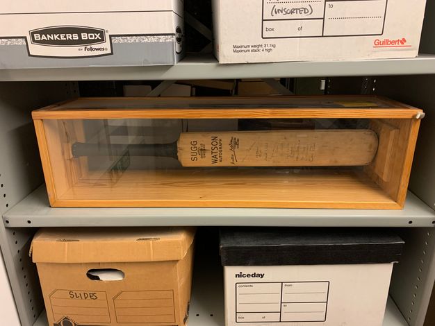 Cricket bat. On a metal storage shelf, a cricket bat is enclosed in a wood and glass case. The manufacturer’s information is near the black handle, reading “Sugg and “Watson Autograph”. A column of about a dozen handwritten signatures along the length of the bat can barely be discerned.