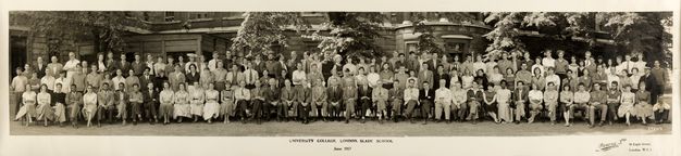 Black and white photograph. Around 125 men in and women, most with pale skin, stand or sit in three long lines in this wide class photograph. The men wear suits or collared shirts, the women dresses, some sleeveless. At the centre of the back row, two men turn to smile at an older man biting down on a pipe. 
