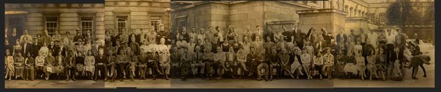 Black and white photograph. Around 125 men in and women stand or sit in three long lines in this class photograph. Many have pale skin but some men and women, the women wearing saris, have darker skin. Some smile, some look serious, and most mug for the camera. One man in a wheelchair holds a rope attached to another man, who pretends to flee to our right.