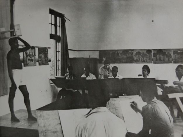 Black and white photograph. A model with dark skin, wearing white shorts, stands with one hand on his head in front of a class. Seven students, all with dark skin, are visible in this image. One is a woman and six appear to be men. A lighter-skinned woman leans over the shoulder of a student across the room from us.