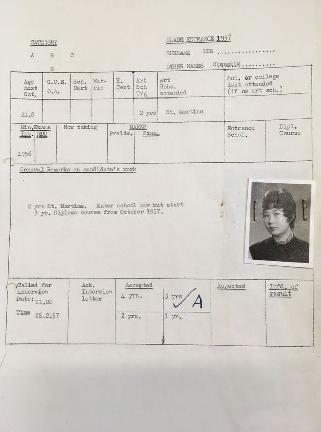 Typewritten form. A passport-sized, black and white photograph of a woman is stapled to a school form. Her short black hair is cut short and sweeps forwards onto her face. She looks at us from the corners of her dark eyes.