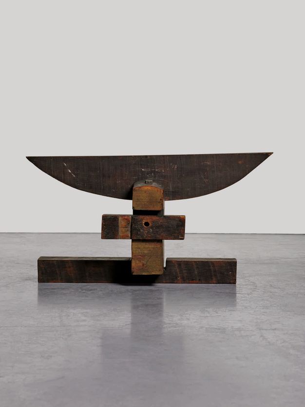 Sculpture. Three wood beams create a base below a flat plank of wood cut into a wide, shallow, cup-like form. At first glance, it could be the silhouette of a wide goblet or a blooming flower.