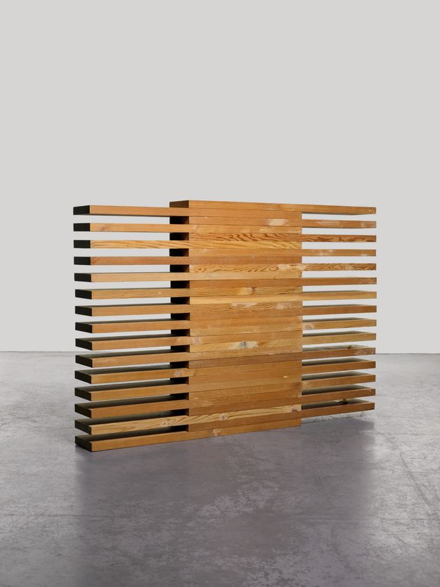 Sculpture. Thirty boards of honey-brown lumber are stacked one atop the other so half of each board overlaps the board below. This creates an alternating, comb-like appearance to each side of the solid stack.