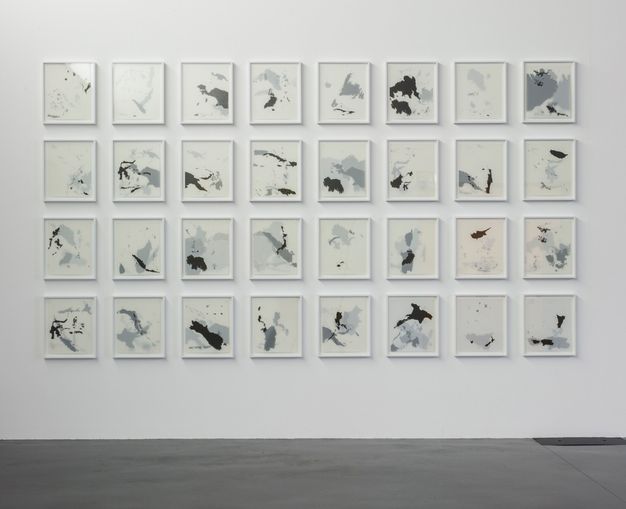 Installation photograph. Grey and white drawings in white frames are hung in a grid of eight rows by four columns. Landmasses silhouetted in grey or black are layered. Some compositions are nearly full and others sparse.